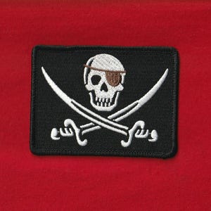 May include: A black rectangular patch with a white skull wearing a brown eye patch, and crossed white swords. The patch is embroidered with white details against a red background. The design is a classic pirate symbol.