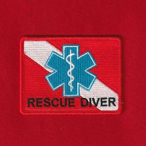 Può includere: Toppa rossa ricamata con un simbolo subacqueo bianco e blu e le parole "RESCUE DIVER" in nero. La toppa è rettangolare con una striscia diagonale bianca.