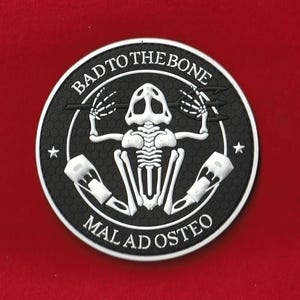May include: A circular black and white patch with the text "BAD TO THE BONE" and "MAL AD OSTEO". The patch features a white skeleton design with a skull, rib cage, and arms holding tridents. Two white stars are on the sides.