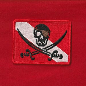 Peut inclure: Un écusson rectangulaire rouge avec un motif de drapeau pirate. L'écusson a une bordure rouge, une bande diagonale blanche et un crâne noir avec un cache-œil. Deux épées croisées sont en dessous.