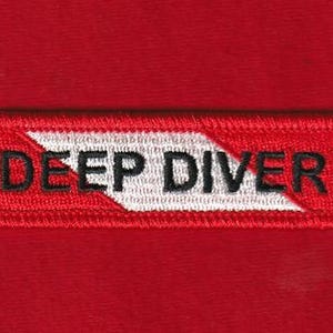 Può includere: Una toppa rettangolare rossa con le parole "DEEP DIVER" ricamate in nero. Un design a chevron bianco divide il testo. La toppa è probabilmente un emblema per gli appassionati di immersioni.