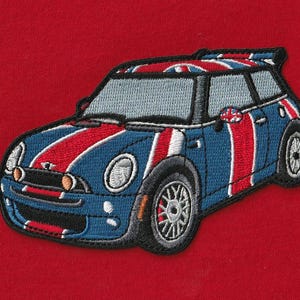 Op de afbeelding: Een geborduurde patch met een blauwe compacte auto met rode en witte racestrepen en een Union Jack-vlagpatroon op het dak. De auto heeft zilveren wielen, een zwarte grille en een kleine achterspoiler, alles tegen een effen rode achtergrond.