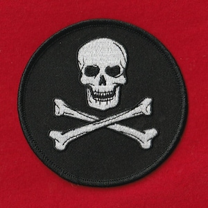 Könnte beinhalten: Ein runder, schwarzer Aufnäher mit einem weißen Totenkopf und gekreuzten Knochen. Der Totenkopf hat detaillierte Augenhöhlen und Zähne. Die gekreuzten Knochen befinden sich unter dem Totenkopf, ein klassisches Piratensymbol.