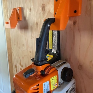 May include: Orange chainsaw wall mount with a black chainsaw handle and a chainsaw. The mount is attached to a wooden wall. The chainsaw is orange and white.