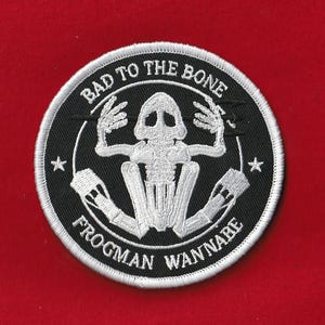 May include: A round, black and white embroidered patch with the text "BAD TO THE BONE" and "FROGMEN WANNABE". The patch features a skeleton frog design in white, with two stars and a white border.