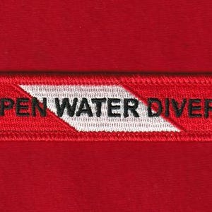 May include: A rectangular red patch with the words "OPEN WATER DIVER" embroidered in black. A white chevron design is in the center of the patch. The patch is on a red background.