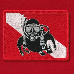 Puede incluir: Parche bordado con un buzo en blanco y negro sobre un fondo rojo. El parche tiene una franja diagonal blanca y un borde rojo. El buzo lleva una máscara y equipo de buceo.