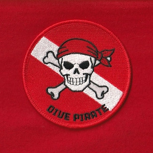 Puede incluir: Un parche redondo y rojo bordado con una calavera blanca con un pañuelo rojo, huesos cruzados y una franja diagonal blanca. El texto "DIVE PIRATE" está bordado en blanco a lo largo del borde inferior del parche.