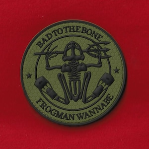 Puede incluir: Parche redondo verde oliva con texto negro y un diseño de hombre rana esqueleto. El texto dice "BAD TO THE BONE" y "FROGMEN WANNABE". El parche tiene un patrón hexagonal texturizado.