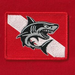 Puede incluir: Parche bordado con un diseño de tiburón. El parche tiene un fondo rojo con una franja diagonal blanca. Un tiburón negro y gris con la boca abierta está centrado en el parche. El parche es rectangular con un borde rojo.