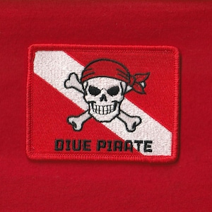 May include: Embroidered patch featuring a skull with a red bandana and crossed bones, set against a red and white background. The text "DIVE PIRATE" is displayed below the skull. The patch is rectangular and has a stitched border.