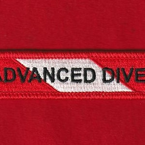 Puede incluir: Un parche rectangular rojo con las palabras "ADVANCED DIVER" bordadas en negro. Un diseño de chevron blanco divide el texto. El parche es probablemente para certificaciones de buceo.