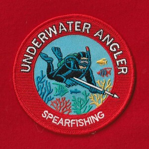 May include: Embroidered patch with a red border featuring a diver spearfishing. The patch has the words "UNDERWATER ANGLER" and "SPEARFISHING" in white. The diver is in a blue circle with fish and coral.