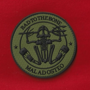 May include: Round, olive green patch with the text "BAD TO THE BONE" and "MAL AD OSTEO" in a circular design. The center features a black skeleton figure holding a trident. The patch has a honeycomb pattern background and two stars.