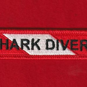 May include: A rectangular red patch with the words "SHARK DIVER" embroidered in black capital letters. A white chevron design is in the center. The patch is on a red fabric background.