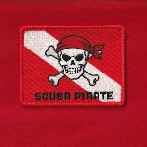 May include: A rectangular patch with a red background features a white diagonal stripe and a skull with crossed bones, wearing a red bandana. The words "SCUBA PIRATE" are embroidered in black below the skull.