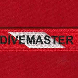 Puede incluir: Un parche rectangular rojo con la palabra "DIVEMASTER" bordada en letras mayúsculas negras. Un diseño de galón blanco está en el lado izquierdo del parche. El parche está sobre un fondo rojo.