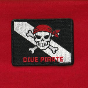 May include: Embroidered patch featuring a skull with a red bandana and crossed bones on a black and white diagonal background. The text "DIVE PIRATE" is in red. The patch is rectangular and set against a red background.