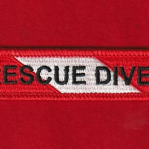 May include: A rectangular red embroidered patch with the words "RESCUE DIVER" in black letters. The patch has a white chevron design. The patch is on a red fabric background.
