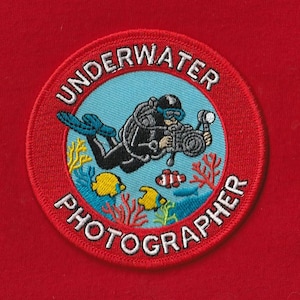 Può includere: Toppa ricamata rotonda con un subacqueo con macchina fotografica, coralli e pesci, con la scritta "UNDERWATER PHOTOGRAPHER". La toppa ha un bordo rosso e un centro azzurro chiaro, su sfondo rosso.