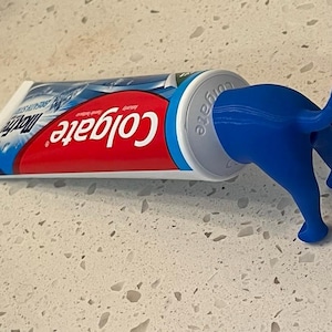 May include: A tube of Colgate toothpaste with a blue dog-shaped toothpaste holder. The toothpaste tube is red and white with blue accents. The dog-shaped holder is blue and is attached to the end of the toothpaste tube.