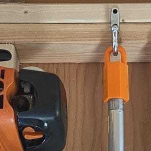 May include: An orange and black power tool is mounted on a wooden wall next to two silver hooks with orange holders. The hooks are designed to hold tools. The wall is made of light brown wood.