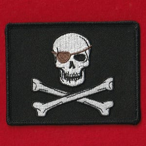 May include: Black rectangular patch featuring a white skull with a brown eye patch and crossed bones. The skull has a detailed design, and the bones are positioned beneath it. The patch is bordered with black stitching and set against a red background.