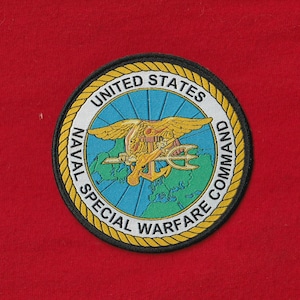 Puede incluir: Parche circular con el emblema del Comando de Guerra Especial Naval de los Estados Unidos. El parche presenta un águila dorada con un tridente, un ancla y un diseño de globo. El texto "UNITED STATES NAVAL SPECIAL WARFARE COMMAND" es blanco.