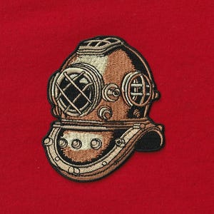 May include: Embroidered patch of a vintage diving helmet in shades of brown, tan, and black, with a detailed design. The patch is set against a solid red background, creating a striking contrast. The helmet features circular viewing ports and intricate metalwork.
