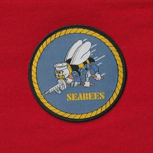 Puede incluir: Parche bordado circular con una abeja de dibujos animados con gorro de marinero y ametralladora. También hay otras abejas de dibujos animados con martillos. El parche tiene un fondo azul con el texto dorado "SEABEES".
