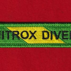 May include: A rectangular embroidered patch featuring a green border and a green background with a diagonal yellow stripe across the centre. The text NITROX DIVER is stitched in black capital letters across the centre of the patch.