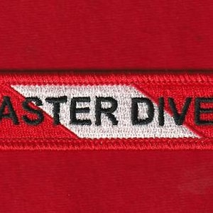 Puede incluir: Un parche rectangular rojo con las palabras "MASTER DIVER" bordadas en negro. El texto está centrado sobre un diseño de galón blanco y negro. El parche tiene un borde rojo.