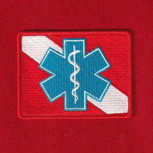 Puede incluir: Parche bordado con un símbolo azul de la Estrella de la Vida y un bastón y una serpiente blancos sobre un fondo rojo. Una franja diagonal blanca cruza el parche. El parche es rectangular con un borde rojo.