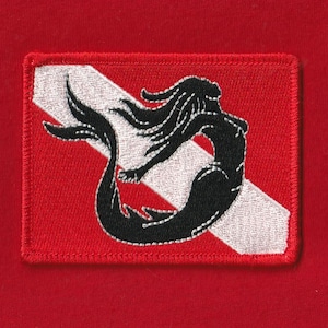 May include: A rectangular red patch with a black mermaid design over a white diagonal stripe. The mermaid has long flowing hair and a fishtail. The patch is embroidered with a red border.