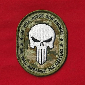 Puede incluir: Un parche bordado con un diseño de calavera. Presenta una calavera blanca con cuencas de ojos negras y un patrón de rayas verticales. El parche tiene un borde verde y el texto "HE WILL JUDGE OUR ENEMIES WE WILL ARRANGE THE MEETING."