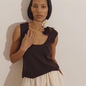Puede incluir: Un top sin mangas marrón oscuro con cuello redondo, combinado con pantalones color crema. Se lleva un collarín blanco con una gran flor blanca. La modelo está de pie frente a una pared lisa de color blanquecino.
