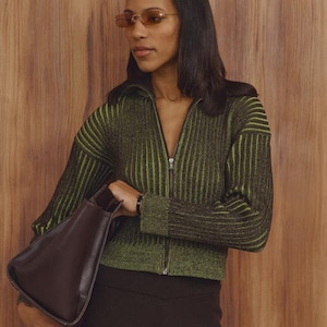 May include: A person wearing a green and black striped zip-up sweater, brown sunglasses, and a dark brown handbag. The sweater has a ribbed texture and a high collar. The handbag is a dark brown, trapezoid shape.