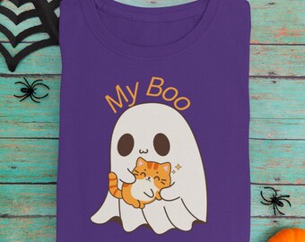 May include: A purple t-shirt featuring a cartoon ghost embracing an orange tabby cat. The text "My Boo" is above the ghost. The shirt is folded on a teal wooden surface, with Halloween decorations.