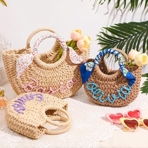May include: Three woven straw bags with round handles. The largest bag features a pink script name and a floral scarf. The medium bag has a blue script name and a blue patterned scarf. The smallest bag displays a purple script name.