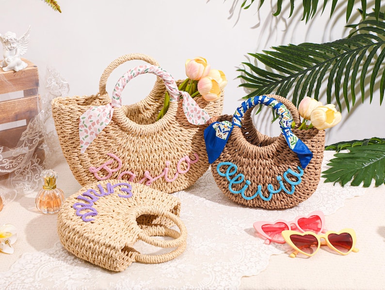 May include: Three woven straw bags with round handles. The largest bag features a pink script name and a floral scarf. The medium bag has a blue script name and a blue patterned scarf. The smallest bag displays a purple script name.