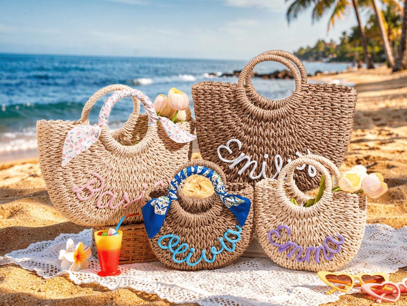 May include: Four woven straw tote bags with round handles, each featuring a custom name in cursive yarn lettering. The bags are decorated with colorful patterned fabric ribbons tied to the handles and include small artificial flowers tucked inside.