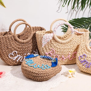 May include: Four woven straw bags with round handles. One dark brown bag features white cursive text, one light tan bag has pink cursive text, one small tan bag has blue cursive text, and one light tan bag has purple cursive text with a floral scarf.