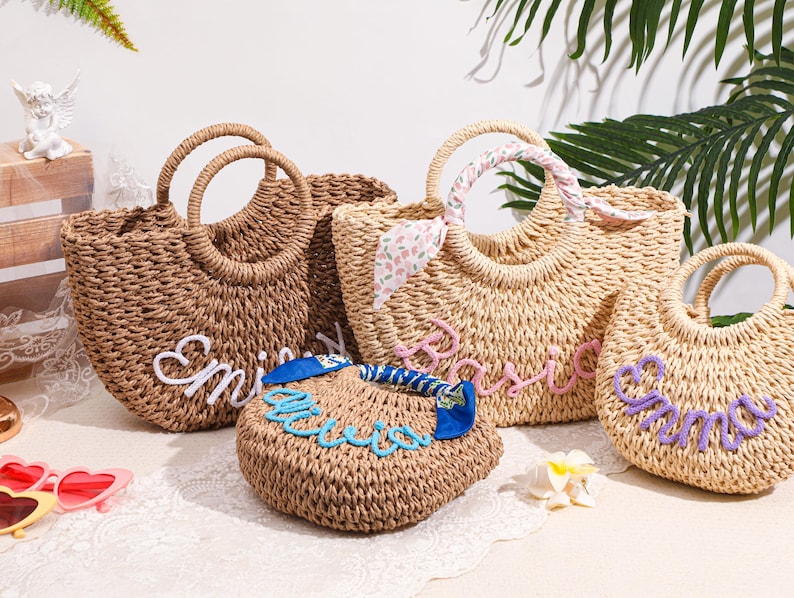 May include: Four woven straw bags with round handles. One dark brown bag features white cursive text, one light tan bag has pink cursive text, one small tan bag has blue cursive text, and one light tan bag has purple cursive text with a floral scarf.