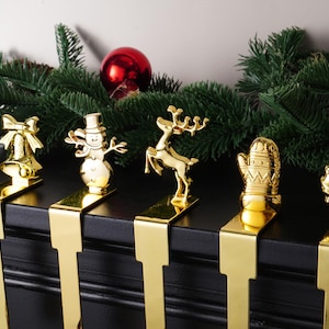 Può includere: Portacalze natalizi dorati a forma di pupazzo di neve, renna e ghirlanda. I supporti sono su una superficie nera, con rami di sempreverdi e una palla rossa sullo sfondo. Decorazioni natalizie.