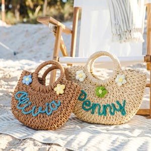 May include: Two woven straw tote bags with round handles. The brown bag features the name Olivia in blue script and two small crochet flowers. The beige bag features the name Penny in blue script and two small crochet flowers in white and green.