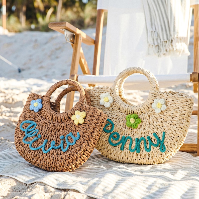 May include: Two woven straw tote bags with round handles. The brown bag features the name Olivia in blue script and two small crochet flowers. The beige bag features the name Penny in blue script and two small crochet flowers in white and green.