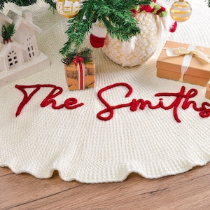 May include: A round, cream-colored knitted Christmas tree skirt featuring the text The Smiths embroidered in bold, red cursive yarn. The textured fabric has a scalloped edge and is displayed beneath a tree with ornaments and gift boxes.