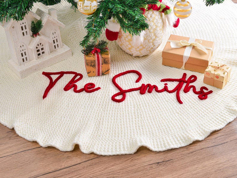 May include: A round, cream-colored knitted Christmas tree skirt featuring the text The Smiths embroidered in bold, red cursive yarn. The textured fabric has a scalloped edge and is displayed beneath a tree with ornaments and gift boxes.