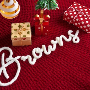 May include: A deep red knitted fabric base features the white cursive text Browns spelled out in thick yarn. The scene includes a gold and white striped ornament, a small gift box with gold dots, a red gift box, and a red gift box with white dots.