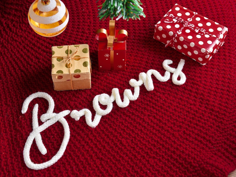 May include: A deep red knitted fabric base features the white cursive text Browns spelled out in thick yarn. The scene includes a gold and white striped ornament, a small gift box with gold dots, a red gift box, and a red gift box with white dots.
